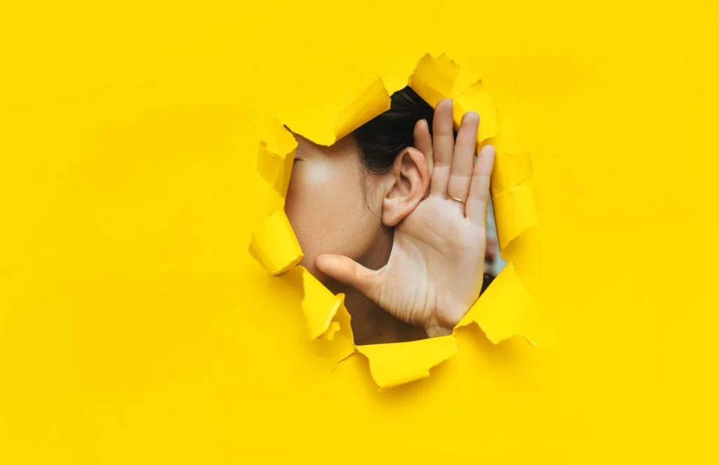 Close-up of a woman's ear and hand through a torn hole in the paper, Yellow background, copy space. The concept of eavesdropping, espionage, gossip and tabloids