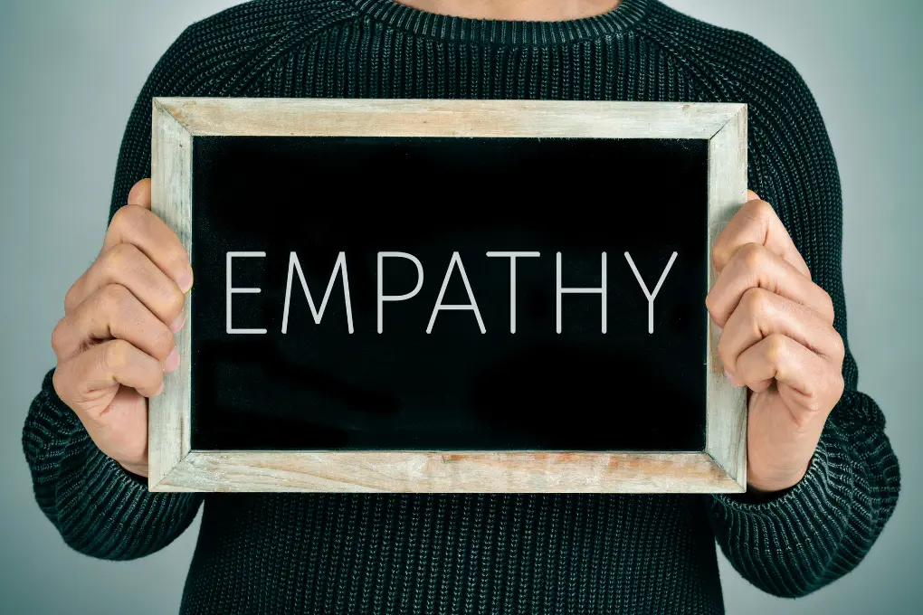 A person wearing a dark sweater holds a small chalkboard with the word EMPATHY written on it in large white letters