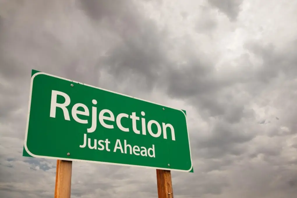 A green road sign reads Rejection Just Ahead against a backdrop of overcast, cloudy skies