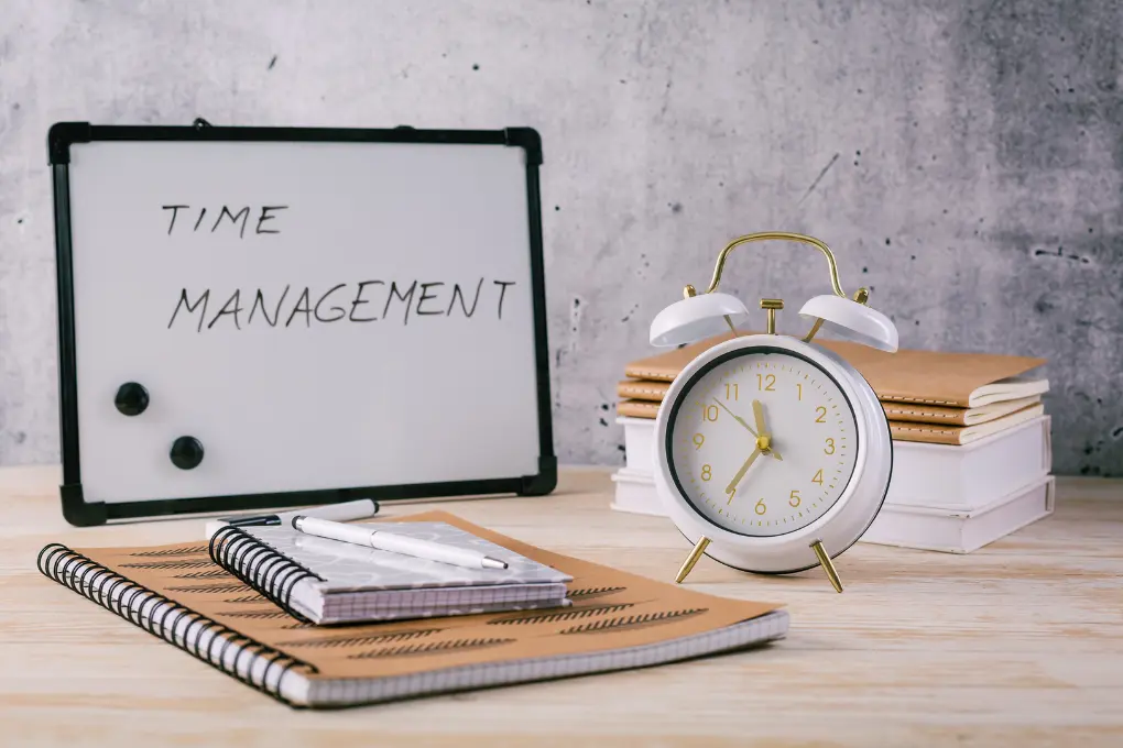 A whiteboard with Time Management written on it is placed on a table alongside a vintage alarm clock, stacked notebooks, and pens