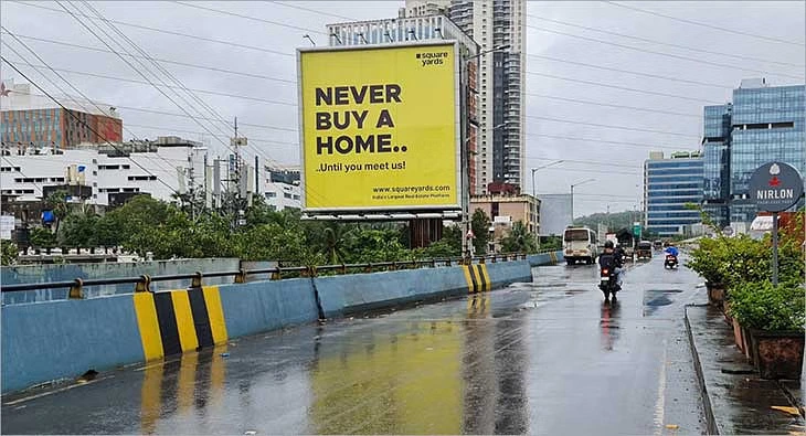 Outdoor billboard advertisement by Square Yards on a rainy day in an urban area, promoting real estate services with the message “Never Buy a Home Until You Meet Us.”